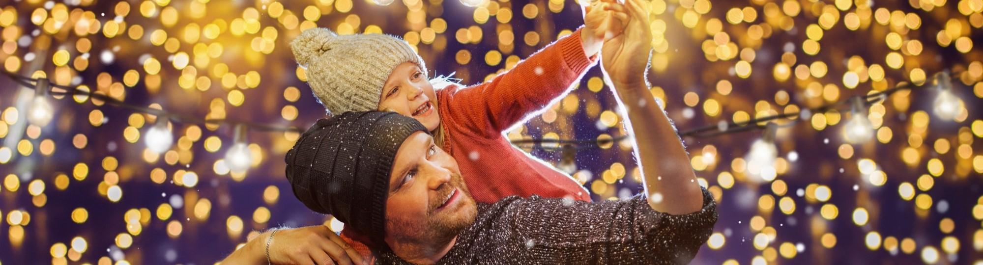 Father giving daughter a piggyback ride while they both reach up toward the Christmas lights.